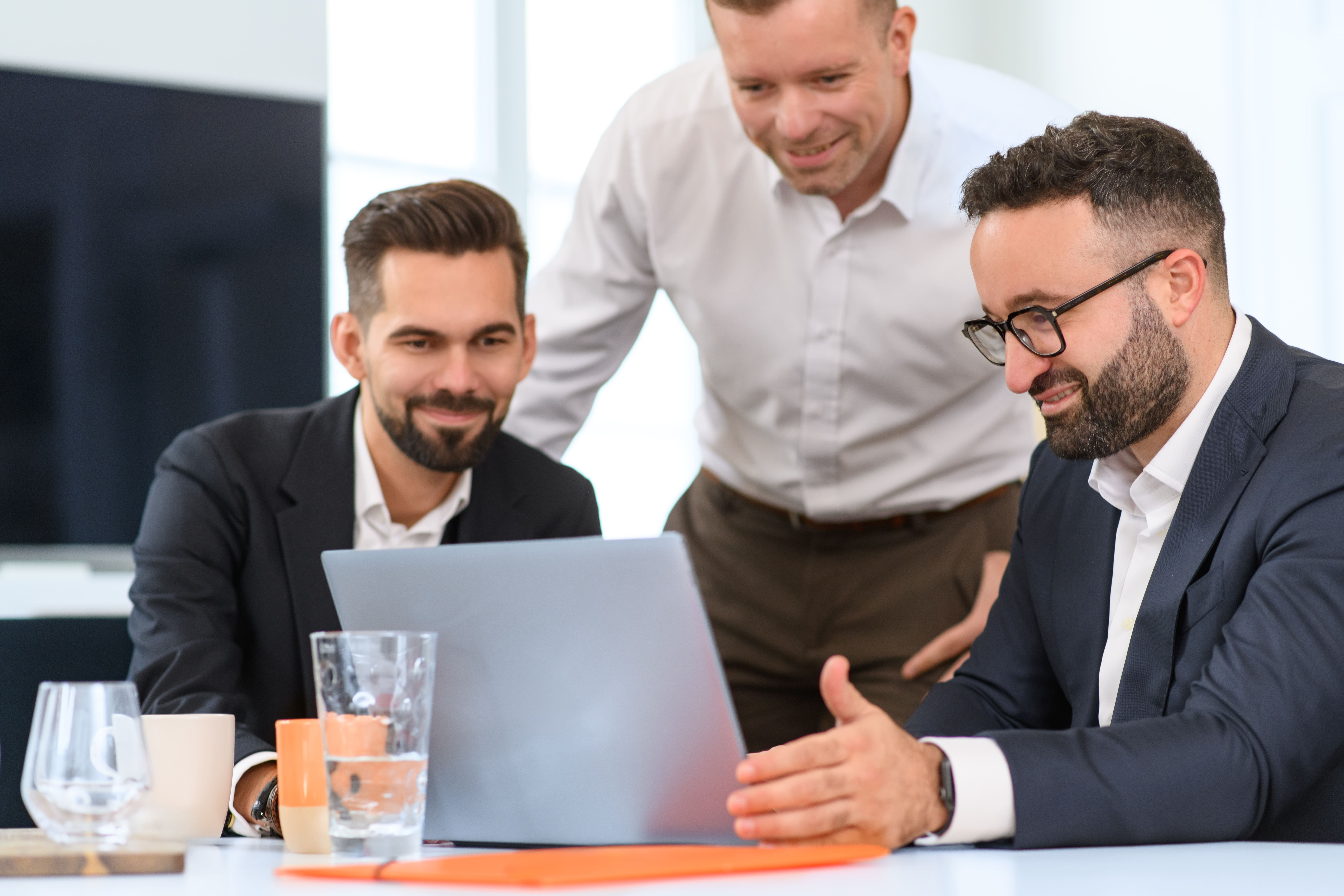 Drei Männer in Businesskleidung stehen und sitzen gemeinsam an einem Tisch und betrachten einen Laptop.