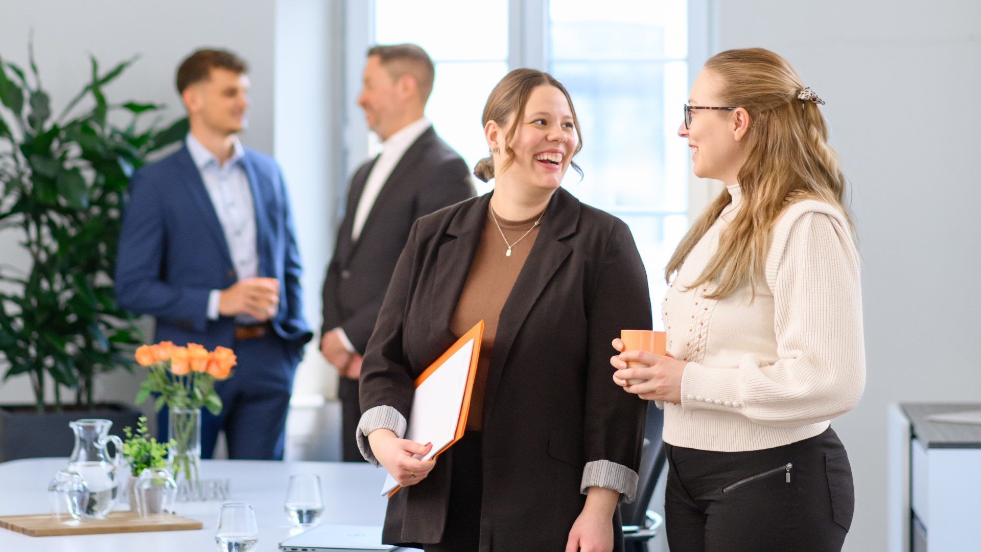 Mehrere Personen im Gespräch in einem hellen Büro, zwei Frauen im Vordergrund unterhalten sich und halten Unterlagen und Tassen in der Hand.